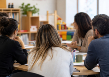 Personas adultas participan en una reunión de formación sobre convivencia infantil alrededor de una mesa, vistas de espaldas