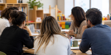 Personas adultas participan en una reunión de formación sobre convivencia infantil alrededor de una mesa, vistas de espaldas